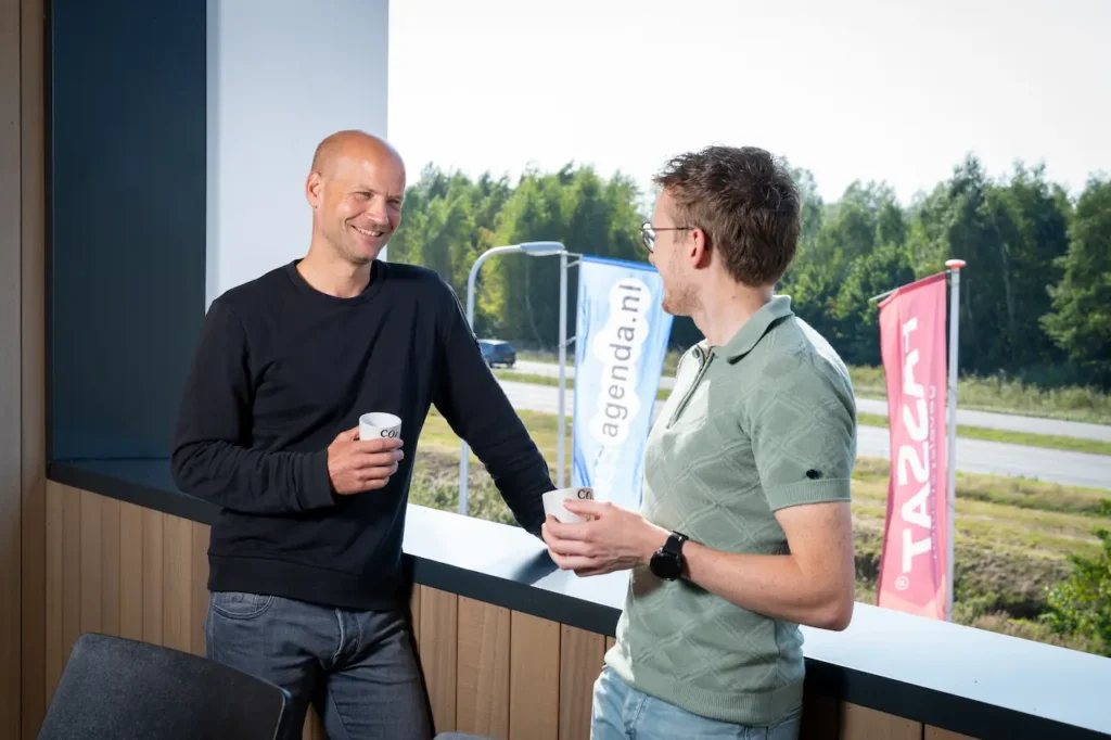 Twee collega's in gesprek op een balkon met vlaggen van Planningsagenda.nl en Tascat