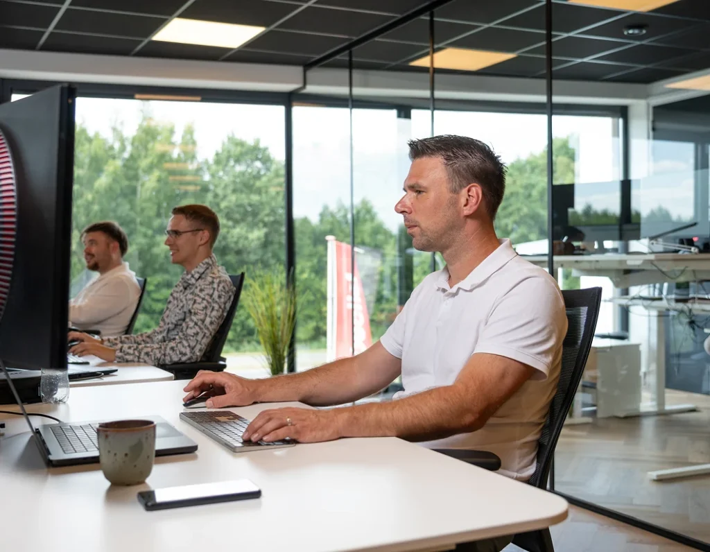 Medewerker werkt achter een beeldscherm, met twee collega's op de achtergrond.