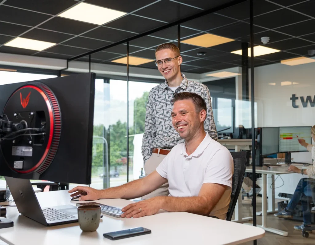 Twee collega's van Twentecs achter een beeldscherm, een van hen glimlacht naar de camera.