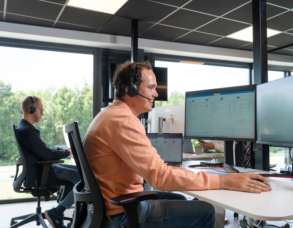 Medewerker van Twentecs met headset, gezien vanaf de zijkant, met collega op de achtergrond